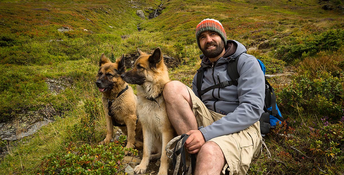 Lächelnder Mann mit Bart, Mütze und Rucksack gewährt sich eine Pause mit seinen zwei Schäferhunden auf einer Bergwiese.